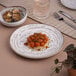 An Ariane white porcelain plate with spaghetti and tomatoes on it with silverware on the table.