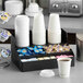 A black plastic coffee condiment organizer with four compartments holding cups, creamers, sugar packets, and stirrers.