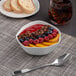 A bowl of fruit and cereal with a silver spoon next to a glass of soda on a table.