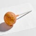 A pumpkin-flavored cake pop coated in orange icing and decorated with yellow and white sprinkles on a stick.
