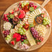 A round wooden charcuterie board featuring assorted cheeses, crackers, fruits, nuts, and meats.