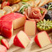 A round wheel of smoked gouda cheese with a red wax coating, partially sliced and displayed on a wooden board with fruit and charcuterie.