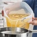 A translucent round 12-quart polypropylene food storage container with blue measurement markings being used to pour liquid through a strainer.