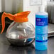 A bottle of Urnex Clearly Coffee 14 oz. liquid coffee pot cleaner next to a glass coffee decanter with an orange handle.