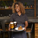 A woman wearing a black short sleeve work shirt and apron, holding a tray with two glasses of beer and a plate of food in a restaurant setting.