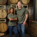 A unisex olive short sleeve work shirt modeled by two people standing in front of wooden barrels.