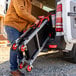 A person loading a foldable three-shelf jobsite cart with red wheels into the back of a van.