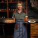 A woman wearing an olive short sleeve work shirt and a denim apron, standing in a bar or restaurant setting while holding trays with food and a drink.