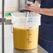 A large translucent round food storage pail with blue measurement markings and a metal bail handle, partially filled with a yellow liquid.