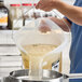 A translucent round food storage pail with a bail handle being used to pour liquid through a strainer.