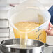 A large translucent round polypropylene food storage container with measurement markings being used to pour liquid through a strainer.