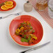 An oval red porcelain plate with a coupe design, featuring a serving of sliced grilled meat garnished with green sauce and citrus zest.