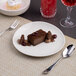 A round, warm white porcelain coupe plate with a slanted edge, shown holding a chocolate dessert.