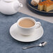 A warm white porcelain tea cup filled with tea, placed on a matching saucer with a spoon nearby.