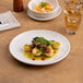 An 8 1/4 inch warm white round porcelain rimless coupe plate with a gourmet food presentation.