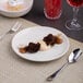 A round, rimless, warm white porcelain coupe plate displayed on a woven placemat with dessert and utensils.