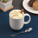 A white porcelain mug filled with a whipped topping and a cinnamon stick, placed next to a spoon.