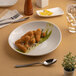 A round, slanted porcelain coupe deep plate in warm white, displayed with food and tableware.