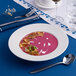 A white round porcelain plate with an embossed wide rim, filled with a vibrant pink soup and garnished with herbs and crispy toppings.