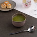 A gold and brown irregular round porcelain bowl filled with green soup, placed on a table with a spoon nearby.
