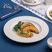 A white round porcelain plate with an embossed wide rim, featuring a serving of roasted vegetables and grains.