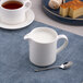 A white porcelain creamer with a handle, filled with milk, placed on a blue placemat next to a spoon.