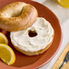 A bagel with a generous layer of light cream cheese, served on a plate with orange slices.