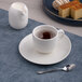 A warm white porcelain coffee cup with saucer, accompanied by a small pitcher of cream, a spoon, and a plate of desserts in the background.