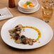 A large, round, warm white porcelain coupe plate with a slanted edge, featuring a gourmet mushroom dish artfully plated.