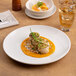 A round, rimless, warm white porcelain coupe plate with a gourmet dish served on it.