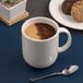 A warm white porcelain coupe mug filled with coffee, shown on a table with a spoon and cookies nearby.