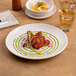 A round, warm white porcelain rimless coupe plate with a modern design, shown with a plated dish.
