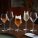 A set of five clear diamond-shaped goblets, one filled with beer, displayed on a wooden table.
