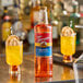 A 1-liter plastic bottle of Torani Orange flavoring syrup placed on a bar counter with two orange drinks garnished with dried citrus slices in the background.
