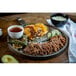 A serving of birria seasoned shredded beef with tortillas, lime wedges, grilled peppers, and a cup of broth on a round platter.