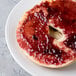 A white porcelain plate holding a bagel half spread with red jam.