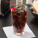 A 12 oz. Libbey Winchester beverage glass filled with a dark soda, ice, and garnished with a cherry, placed on a white napkin.