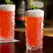 A textured clear glass filled with a red cocktail, topped with foam and garnished with berries and a mint sprig.