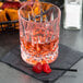 A clear, faceted 10 oz. rocks glass with a reddish beverage, placed on a dark napkin with two maraschino cherries on a cocktail pick.