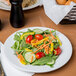 A white porcelain plate with a fresh salad containing arugula, grape tomatoes, cucumber slices, shredded carrots, and yellow bell pepper strips.