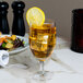 A clear glass goblet filled with an amber beverage, ice cubes, and garnished with a lemon slice on the rim.