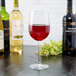 A clear 16 oz. wine glass filled with red wine, surrounded by bottles of wine on a dark surface.