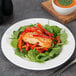 A white wide rim porcelain plate with a serving of salmon, sautÃ©ed red peppers, and fresh greens.