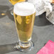 A clear 12 oz. footed pilsner glass filled with light beer and topped with foam.