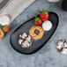 A black, oval-shaped porcelain tray displaying assorted cookies and strawberries.