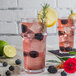 A clear, stackable 16 oz. plastic cooler glass filled with a pink beverage, ice, blackberries, and garnished with a lemon slice and rosemary sprig.