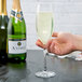 A clear 8 oz. champagne flute filled with sparkling wine, held by a hand, with bottles of champagne in the background.