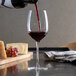 A clear wine glass with pour control markings being filled with red wine, placed on a table next to cheese and grapes.