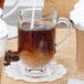 A clear glass coffee mug with a handle, partially filled with coffee and cream, placed on a lace doily.