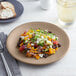 A tan matte porcelain coupe plate with a colorful salad served on it.